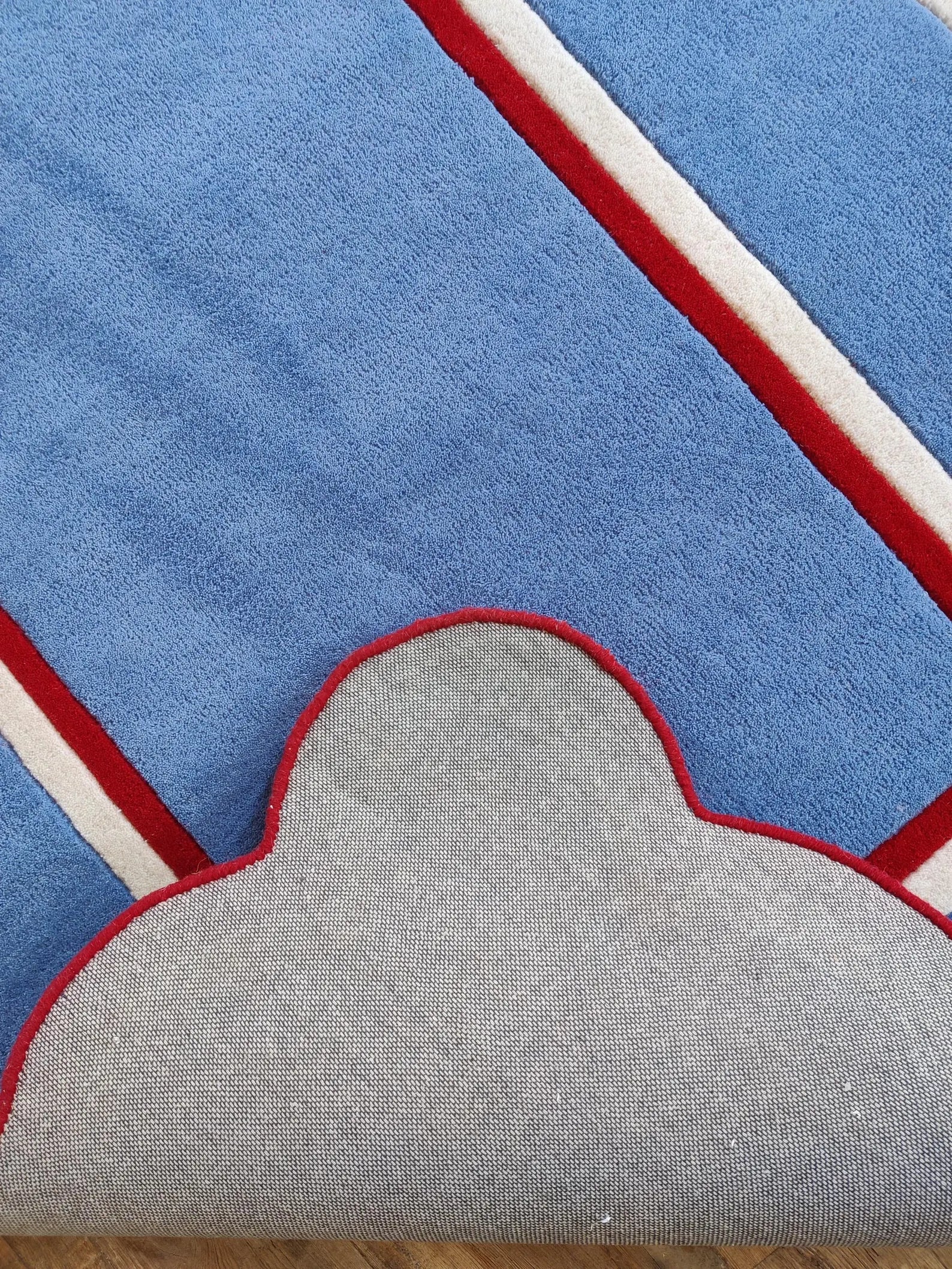 Close-up of a blue textured rug with diagonal red and white stripes and a scalloped gray underside, showing the pile direction and edge trim.
