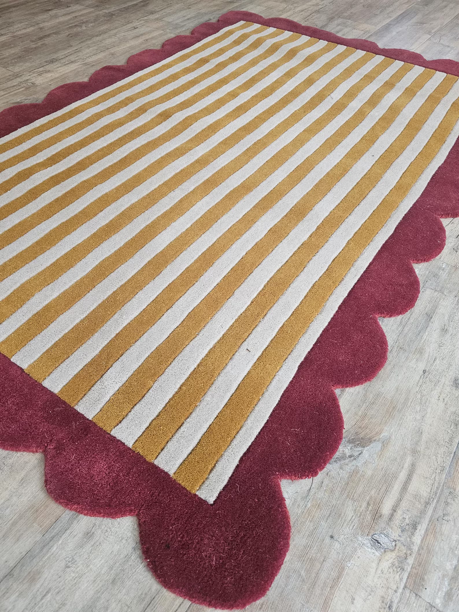 A rectangular area rug with mustard yellow and off-white vertical stripes and a scalloped burgundy border, shown on a light wood floor.
