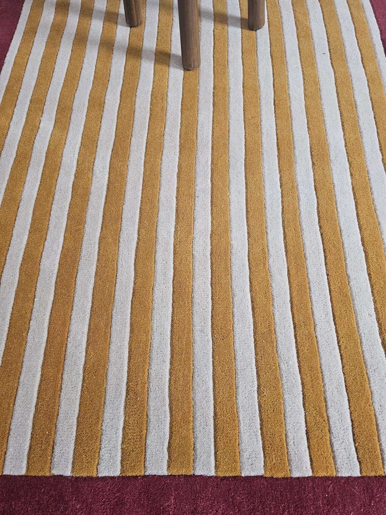 A close-up of a striped area rug with alternating mustard yellow and off-white vertical stripes, shown with the legs of a wooden chair visible at the top.