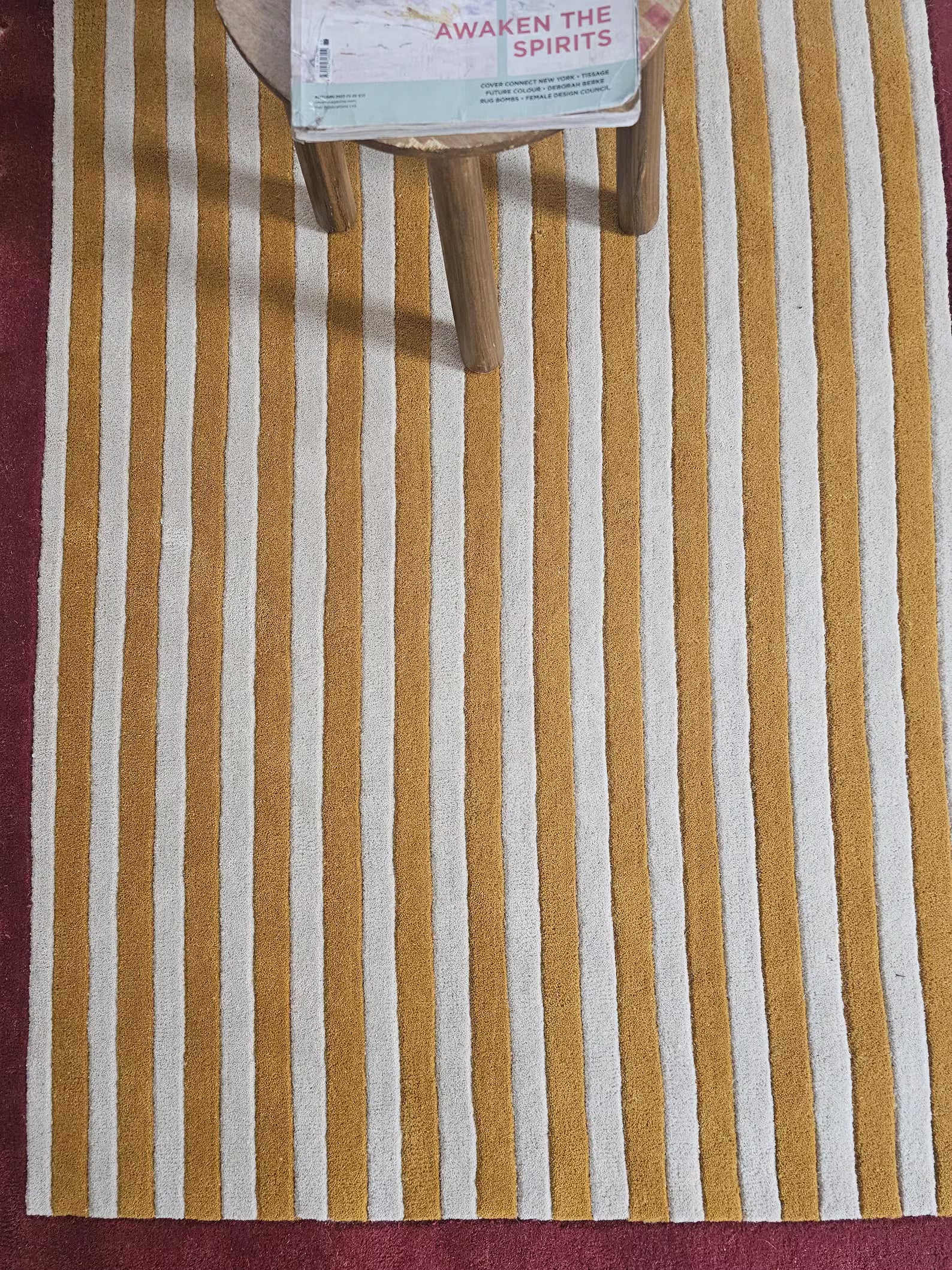 A mustard yellow and off-white vertically striped rug laid on a dark red floor, with the lower legs of a wooden stool visible at the top edge.