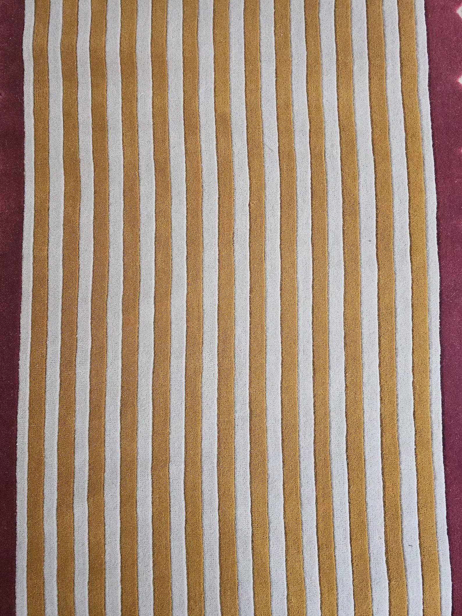 A close-up of a striped rug pattern with alternating vertical bands of mustard yellow and off-white, bordered by a dusty burgundy edge.
