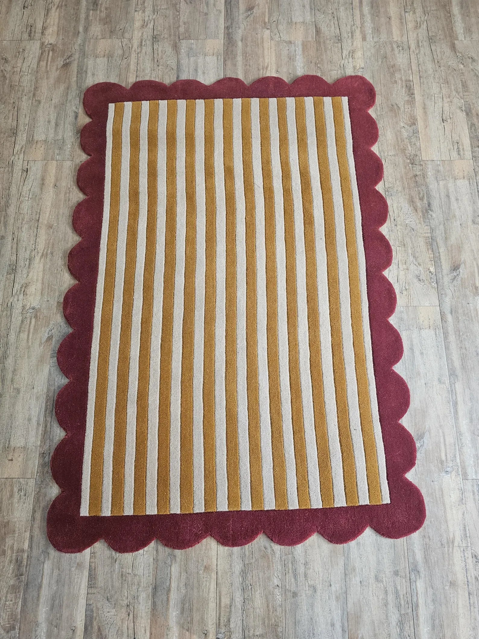 A rectangular area rug with vertical mustard yellow and cream stripes, finished with a scalloped dark red border, shown from a top-down view on a light wood floor.