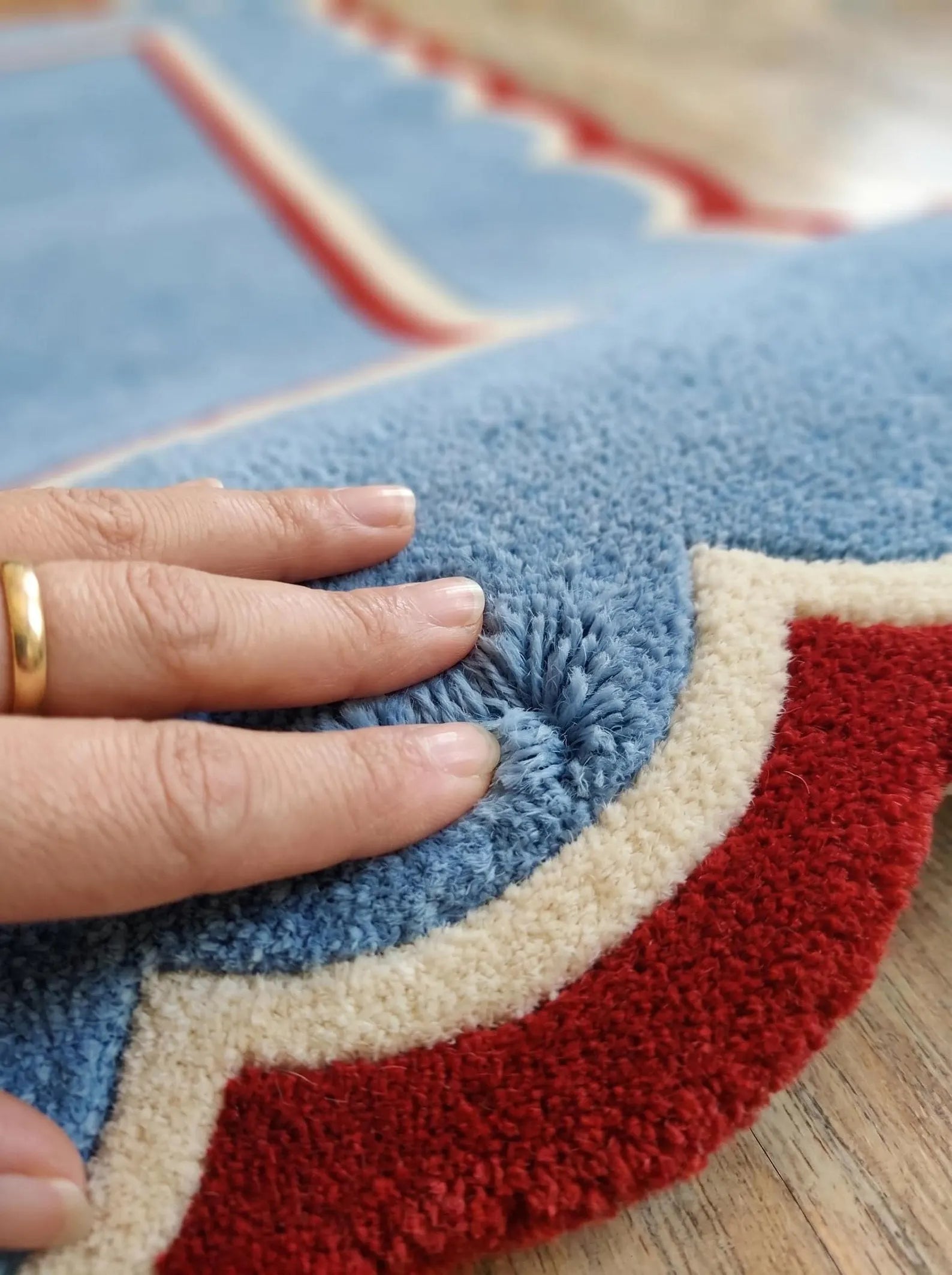 A close-up of a hand pressing down on the textured loop pile of a blue, red, and cream patterned rug on a wooden floor.