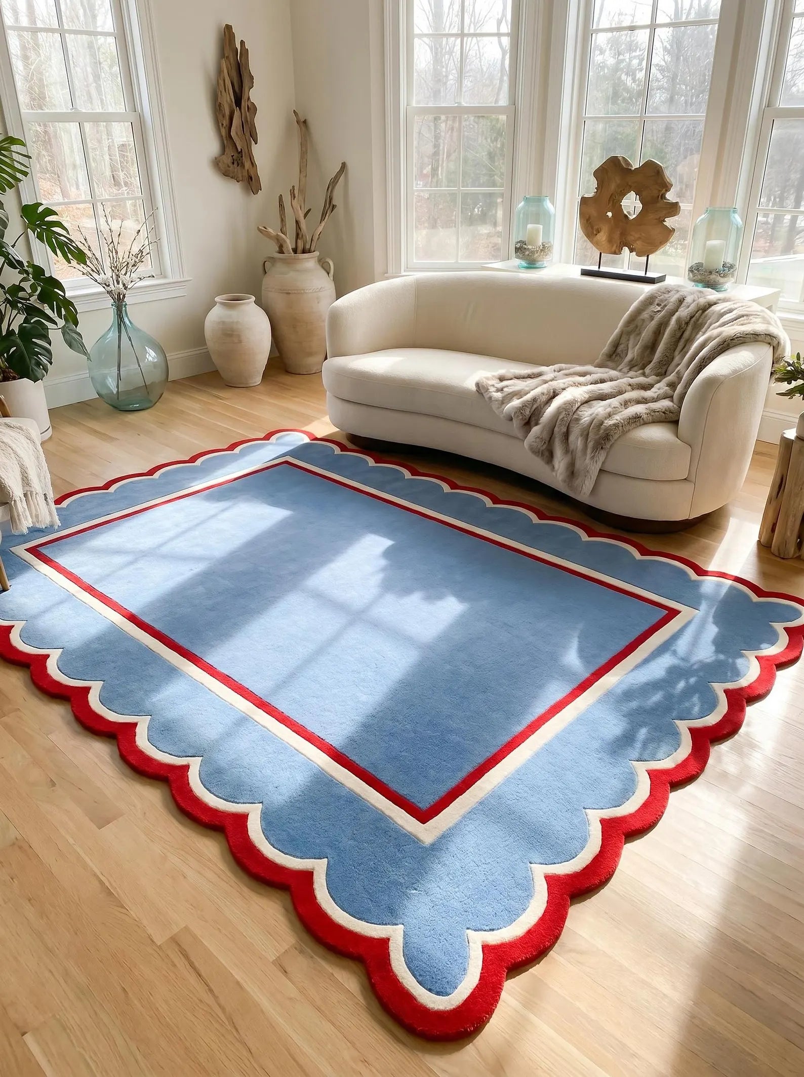 A rectangular area rug with a scalloped edge, featuring a light blue center, a red inner border, and a cream outer border. The rug is styled in a bright, modern living room with a curved white sofa, wooden floors, and decorative vases.