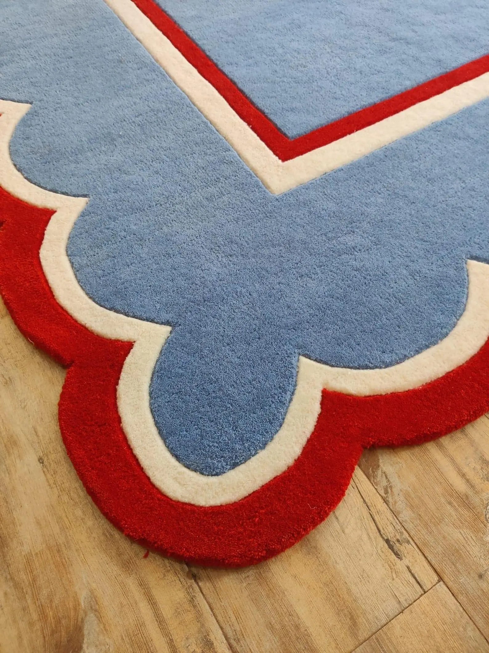 A close-up of a plush area rug with a scalloped border, featuring a light blue center, a red outer border, and a cream inner border, shown on a light wood floor.