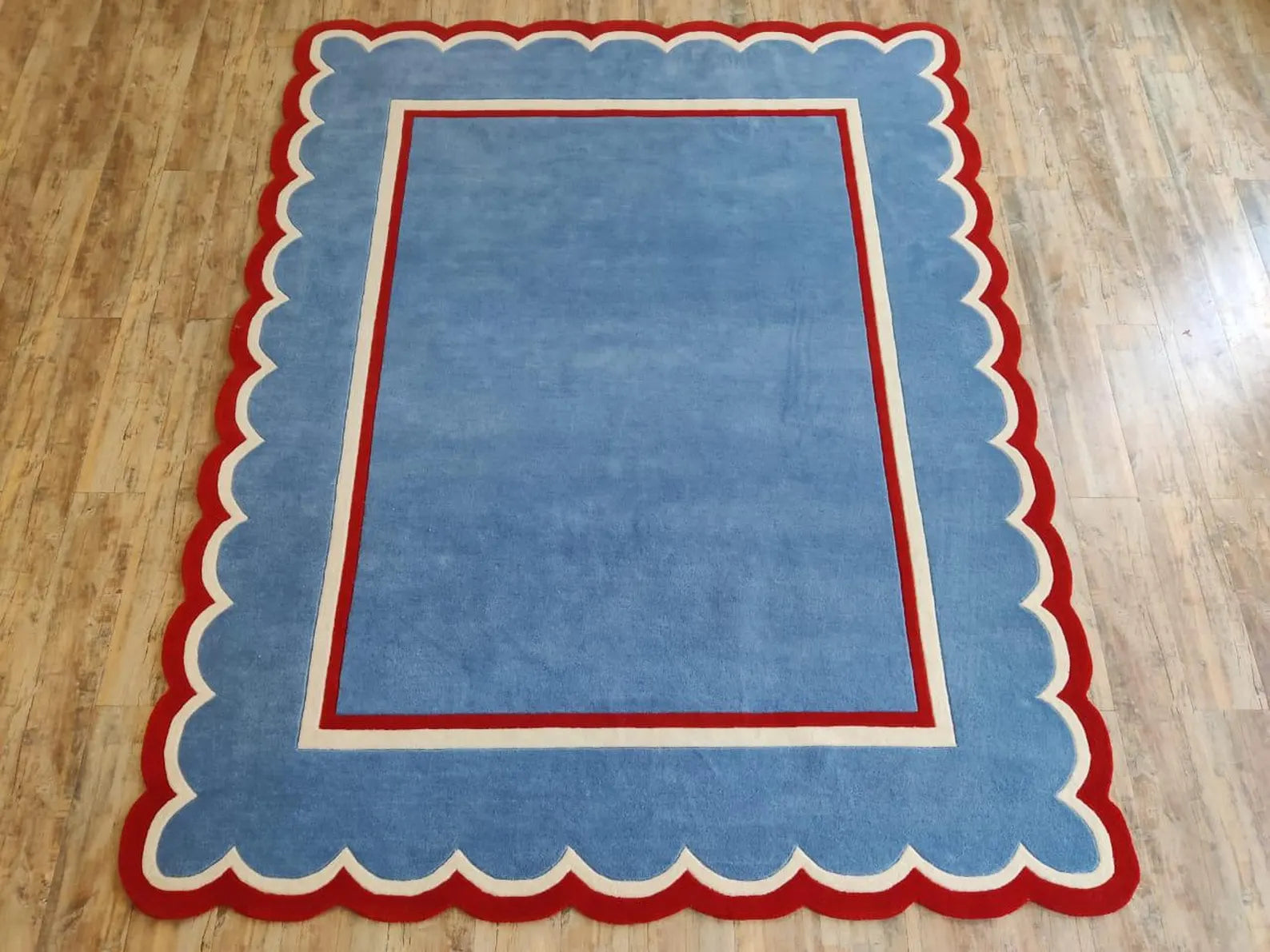 A rectangular area rug with a scalloped border, featuring a light blue center, a red inner border, and an outer red and cream scalloped edge, displayed on a light wood floor.