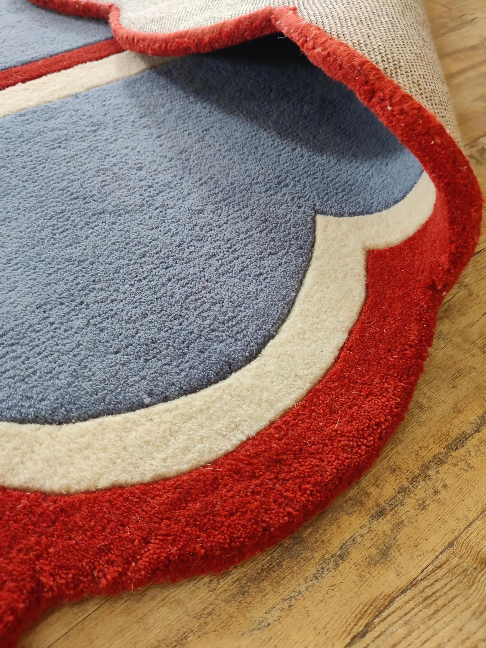 A close-up of a plush area rug with a layered abstract design in slate blue, cream, and red, shown partially folded over on a light wood floor.