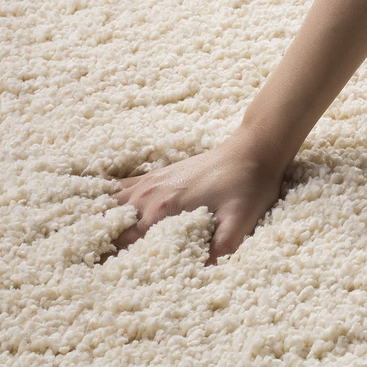 A close-up of a person's hand pressing into a thick, plush, off-white shag rug to demonstrate its soft texture and depth.