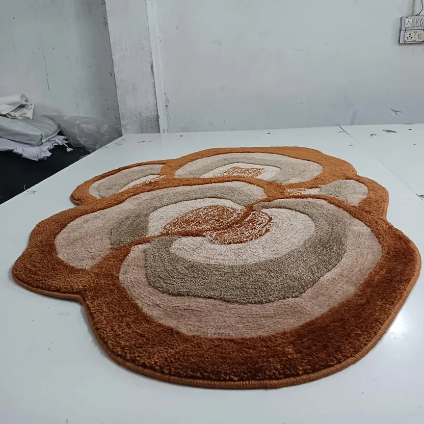 A plush, multi-tiered circular rug with an abstract, concentric petal-like design in shades of rust brown, tan, and cream, displayed on a white table.