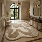 A large abstract area rug in cream and taupe tones is displayed in a modern, elegant entryway with a console table, wall mirror, and arched glass doors in the background.