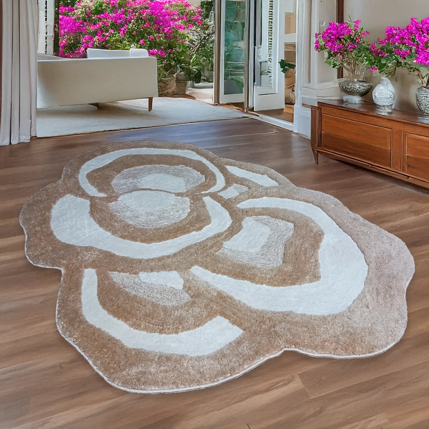 A large, organic-shaped shag rug with a raised floral or abstract petal pattern in taupe brown and off-white, displayed on a dark wood floor in a bright, modern living room.