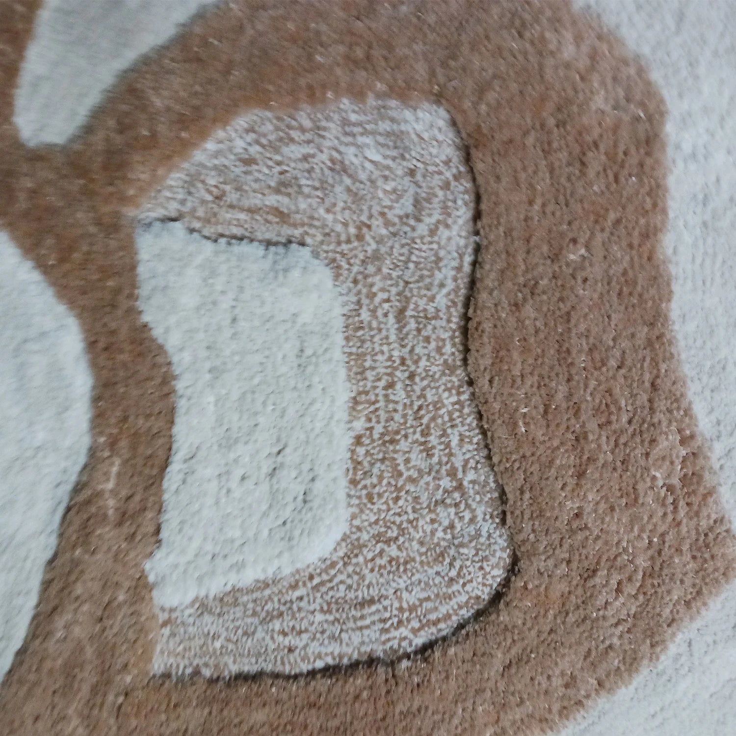 Close-up of a plush tufted rug with a raised abstract shape in warm taupe-brown and off-white.
