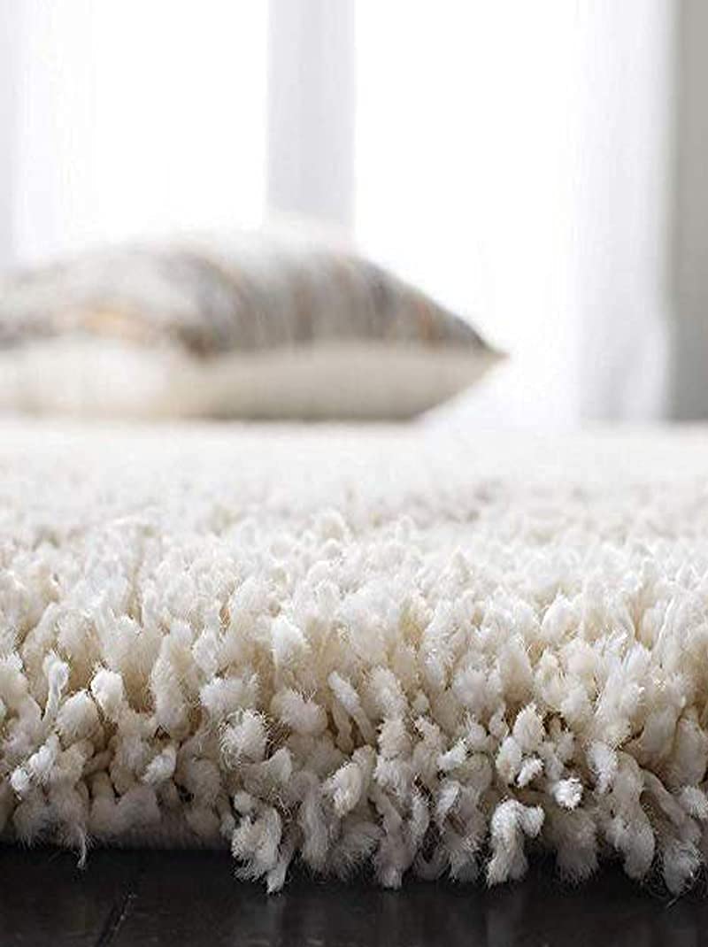 A close-up of a plush, high-pile shag rug in off-white or cream, with a blurred decorative pillow in the background near a bright window.