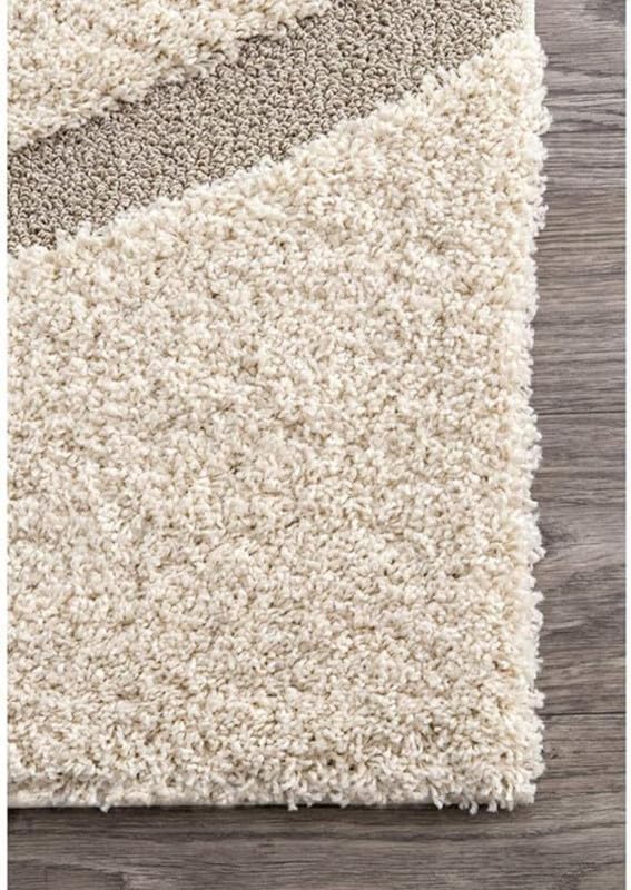 Close-up of a shag area rug corner showing a thick cream pile with a diagonal taupe band, placed on a gray wood floor.