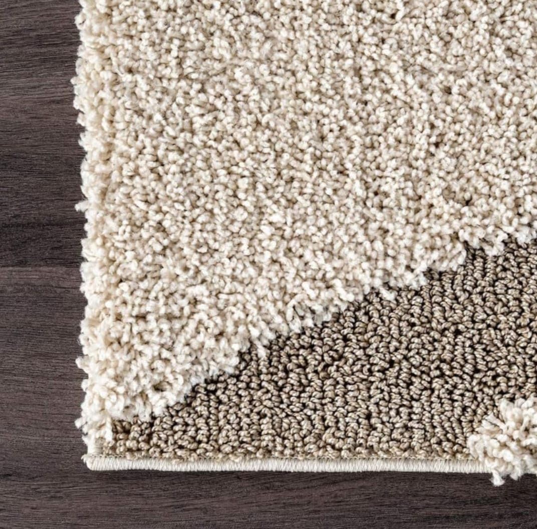 Close-up of a shag area rug corner showing a thick cream pile on top and a taupe-beige underside, placed on a dark wood floor.