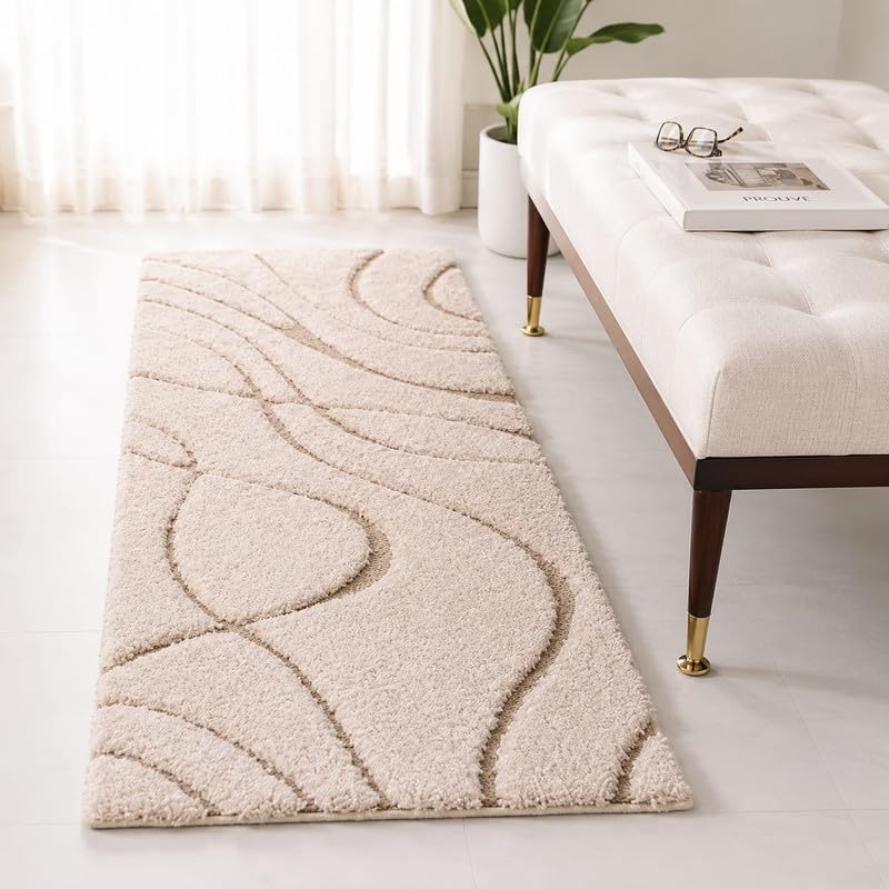 A rectangular shag area rug in a warm cream or light beige color with a raised wavy abstract pattern, styled in a bright room next to a tufted bench.