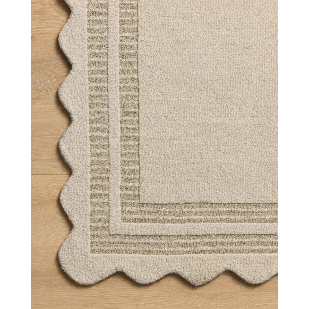 Close-up of a cream-colored rug corner with a scalloped edge and raised striped border, shown on a light wood floor.