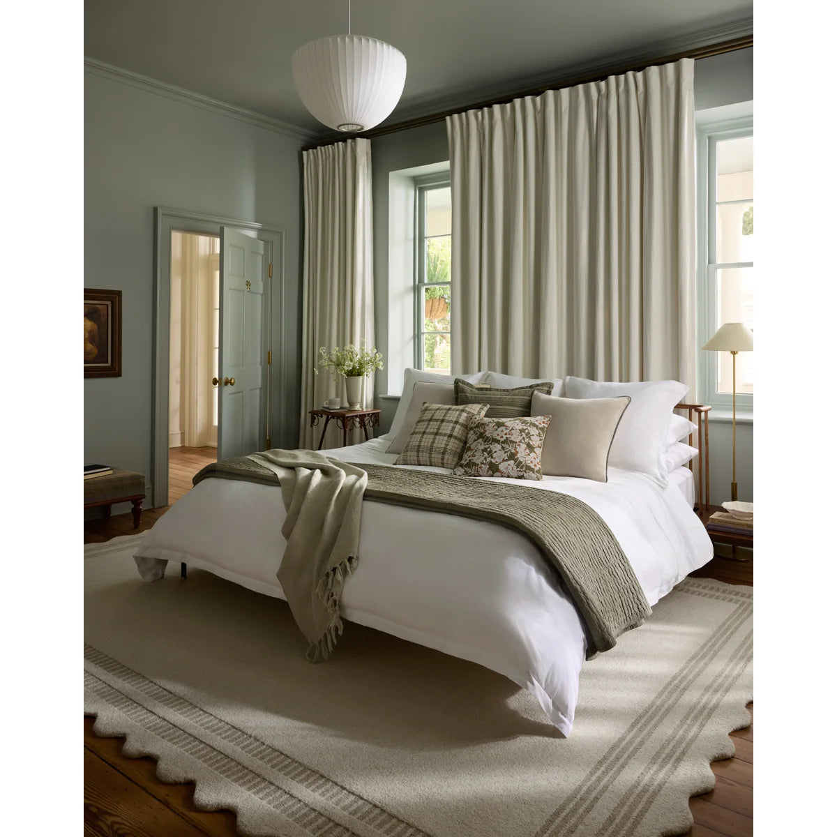 A styled bedroom interior featuring a bed dressed in white linens with neutral-toned pillows and a textured throw blanket, set in a room with beige curtains, a patterned rug, and soft natural light from windows.