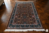 Naqsh Persian rug flat lay on wooden floor — all-over floral arabesque in warm brown with deep teal border and ivory fringe