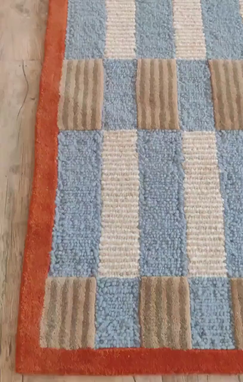 Powder blue, cream, and tan woven check bordered by a solid rust edge — a tonal field with a warm contrast frame rug 