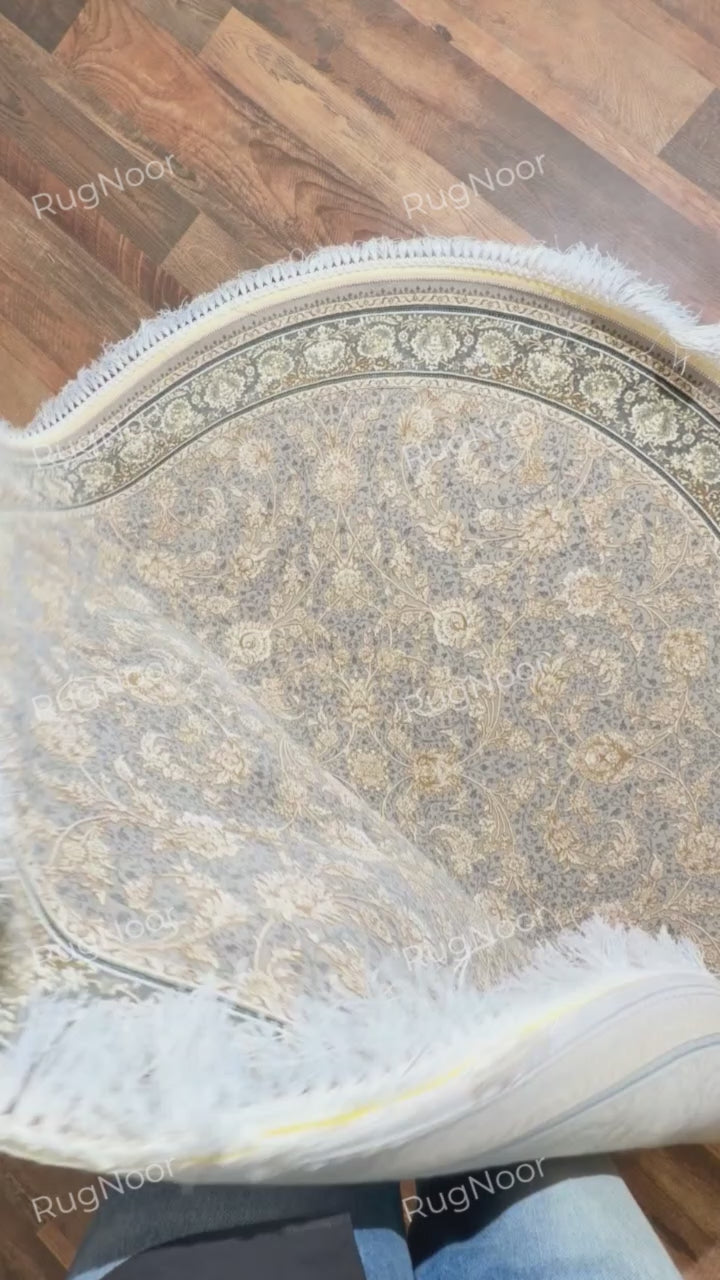 Golnaré grey round carpet with a corner folded back, revealing the pale underside alongside the floral pattern and ivory fringe.