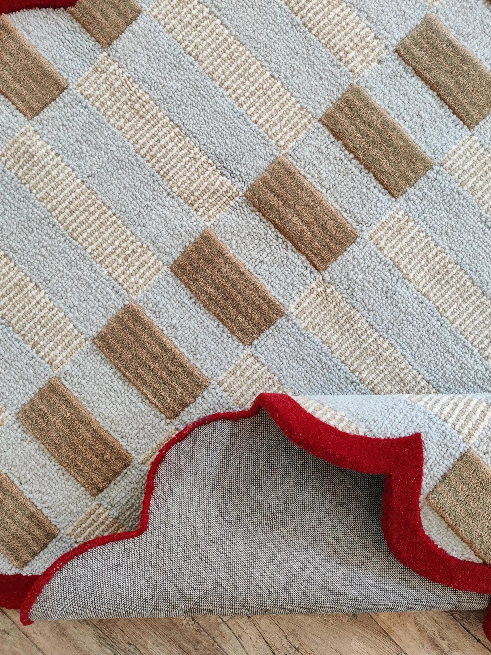 A close-up of a textured area rug with a geometric pattern of beige, brown, and light gray woven blocks, shown with one corner folded over to reveal a red scalloped edge.