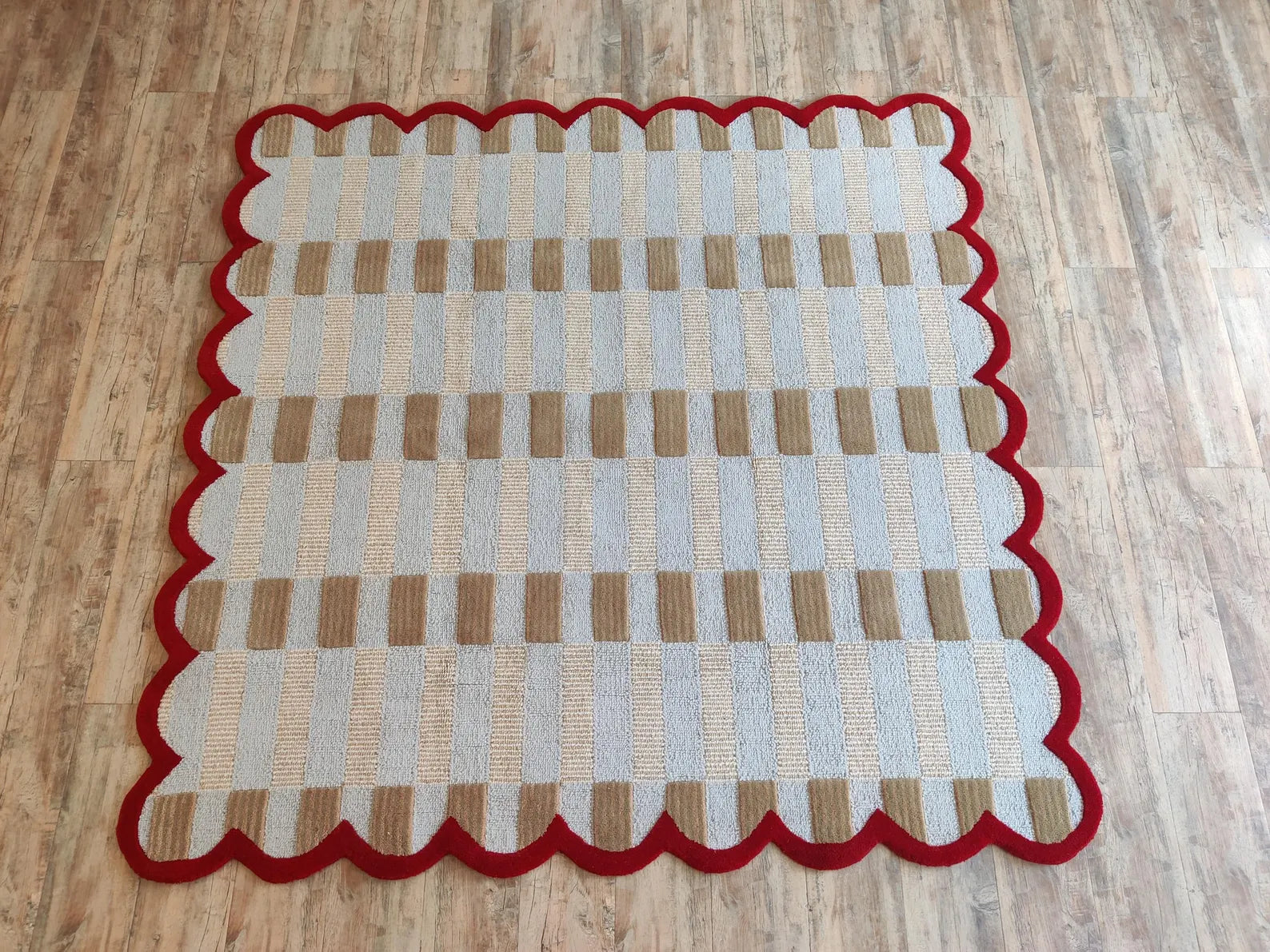 A square area rug with a scalloped red border, featuring a woven pattern of beige, light gray, and brown stripes, shown on a light brown wood floor.