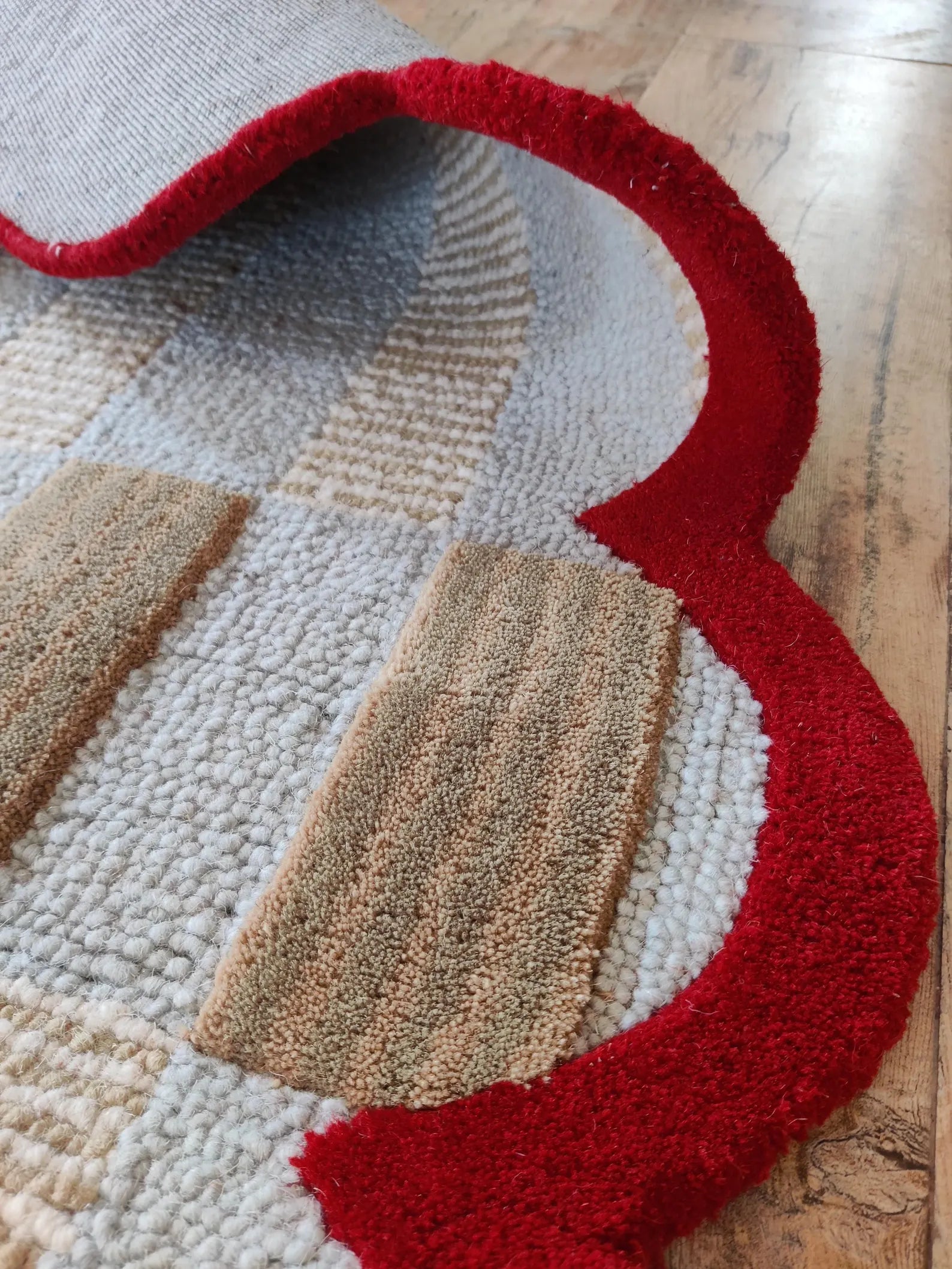 A close-up of a textured area rug with a bold red border, beige and off-white checkered pattern, and chunky looped pile, shown partially rolled up on a wooden floor.