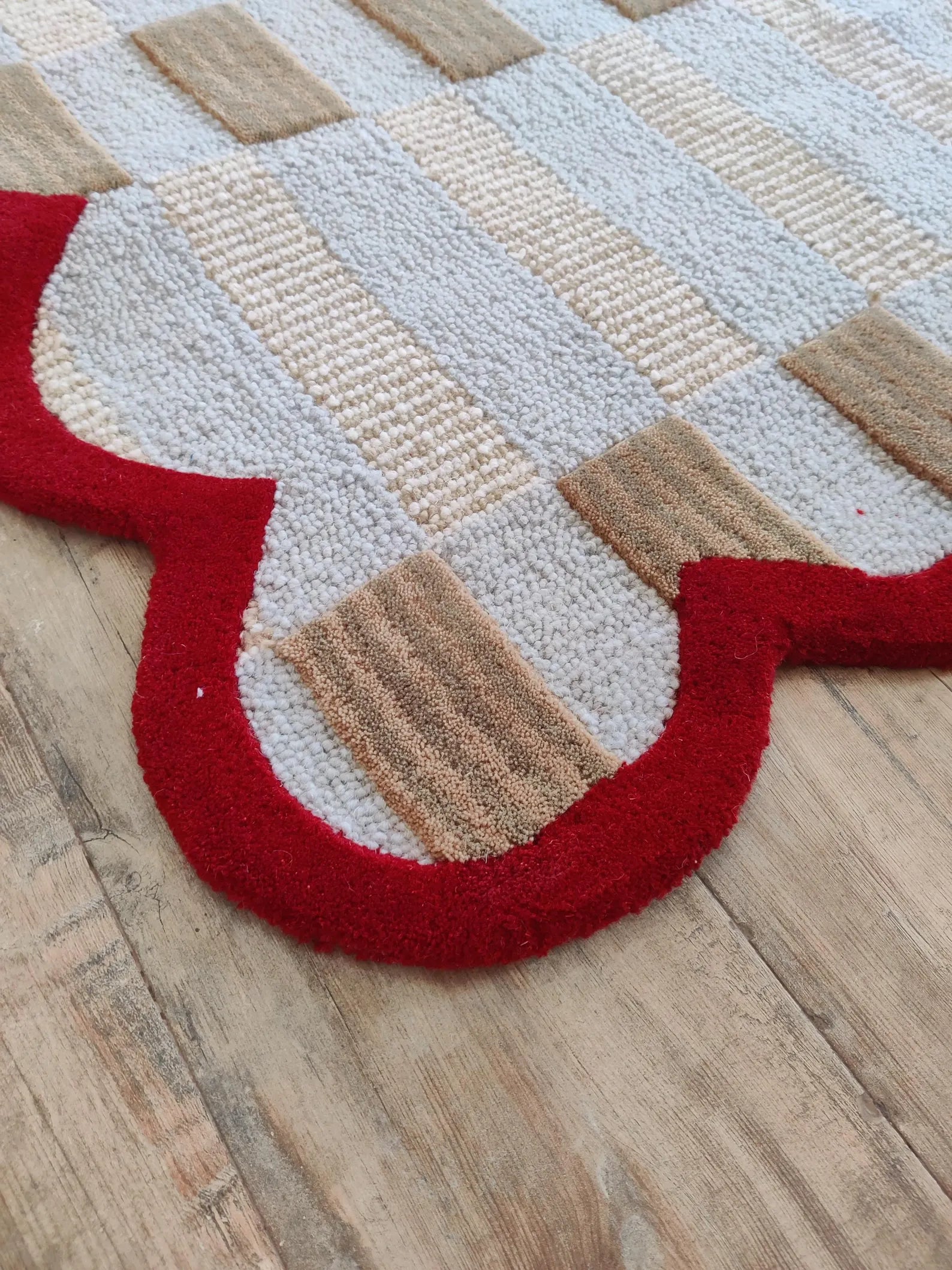A close-up of a textured area rug with a scalloped border, featuring a patchwork design of beige, cream, and red sections, laid out on a light wood floor.