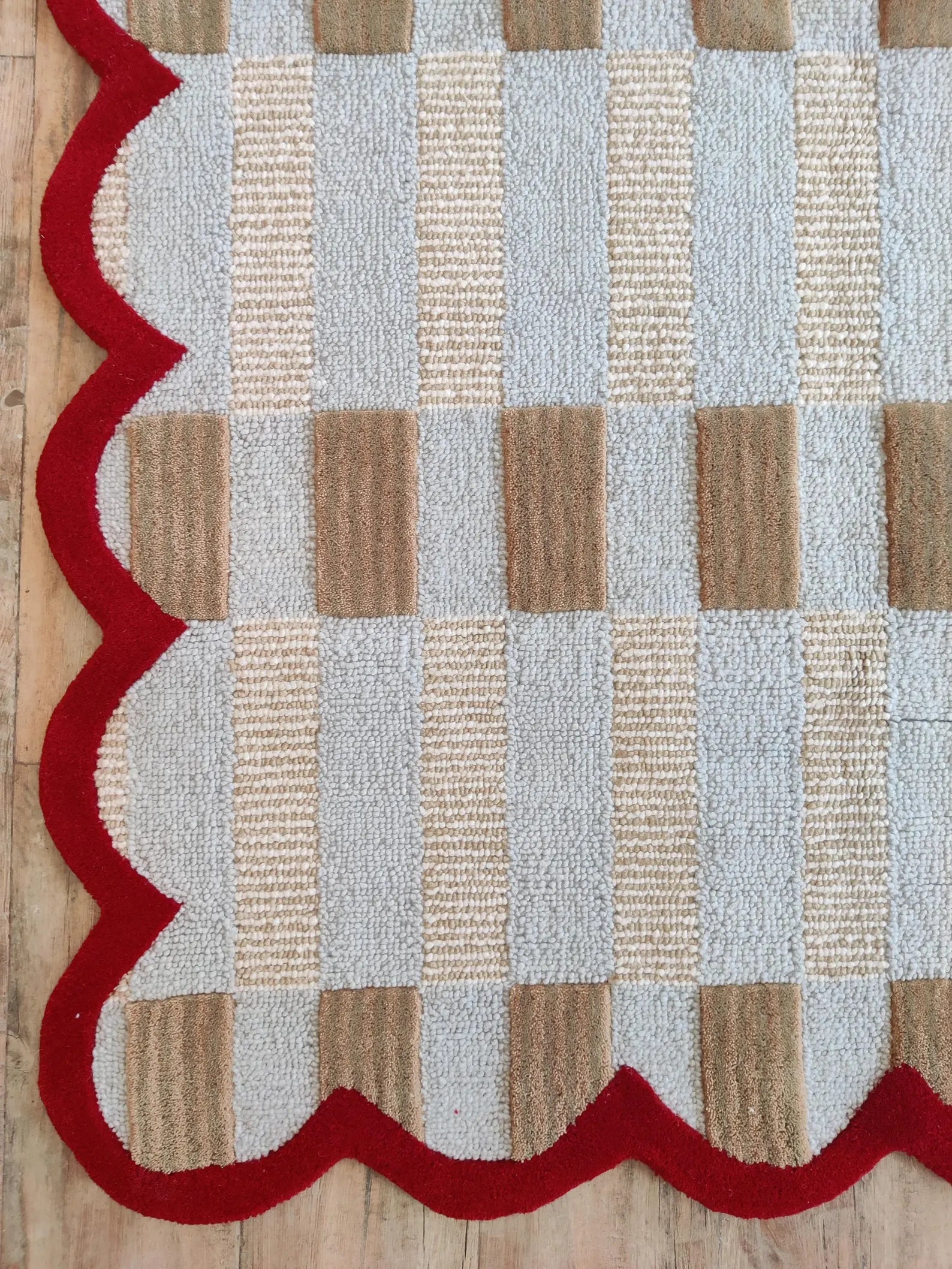 A close-up of a woven rug with a scalloped edge, featuring a checkered pattern of beige, light blue, and brown squares with a bold red border, laid on a light wood floor.