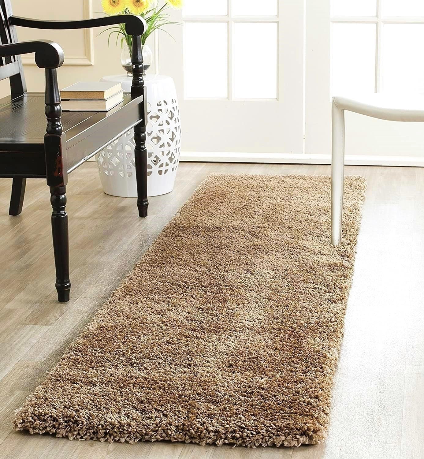 A long, low-pile shag runner rug in a warm taupe-brown color, styled on a light wood floor in a bright, modern room next to a dark wooden chair and a white stool.