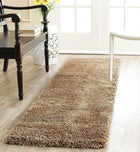 A long, low-pile shag runner rug in a warm taupe-brown color, styled on a light wood floor in a bright, modern room next to a dark wooden chair and a white stool.