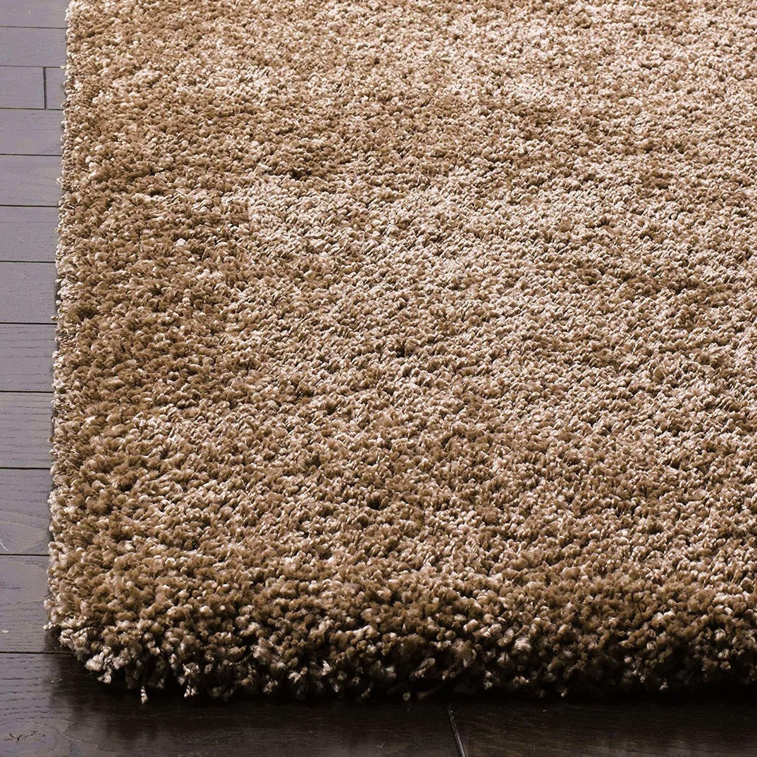 A close-up of a shag area rug in a warm taupe-brown and beige colorway, shown resting on a dark wood floor.