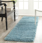 A long, rectangular shag runner rug in a dusty light blue color, shown on a light wood floor in a bright, modern room setting.