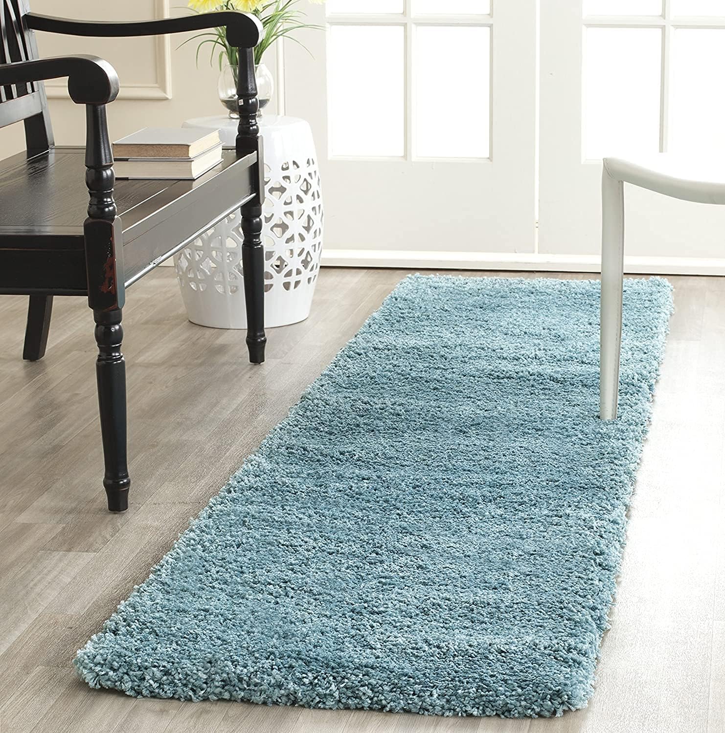 A long, rectangular shag runner rug in a dusty light blue color, shown on a light wood floor in a bright, modern room setting.