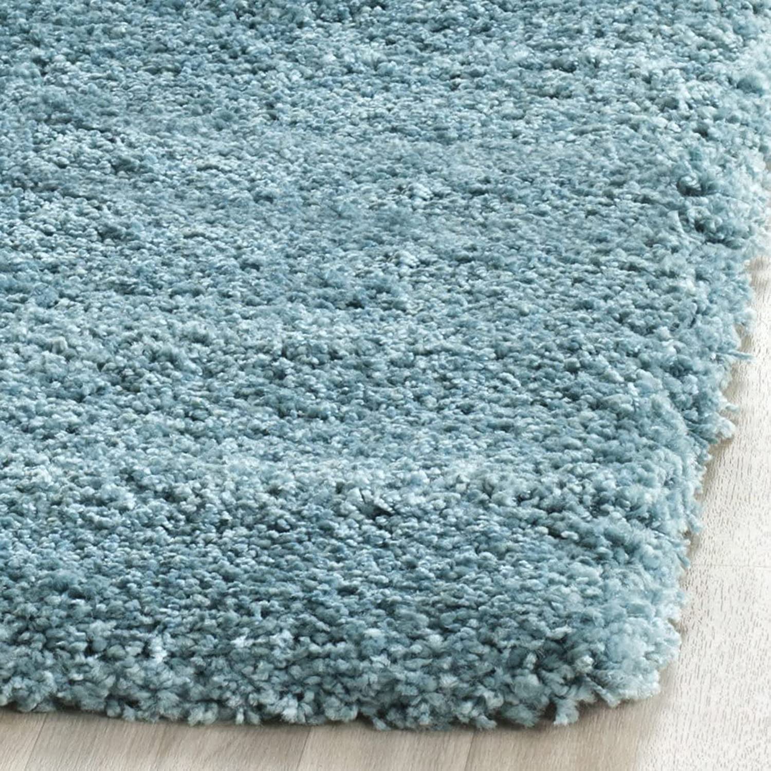 A close-up of a plush, high-pile shag rug in a dusty light blue color, shown resting on a light-colored wooden floor.