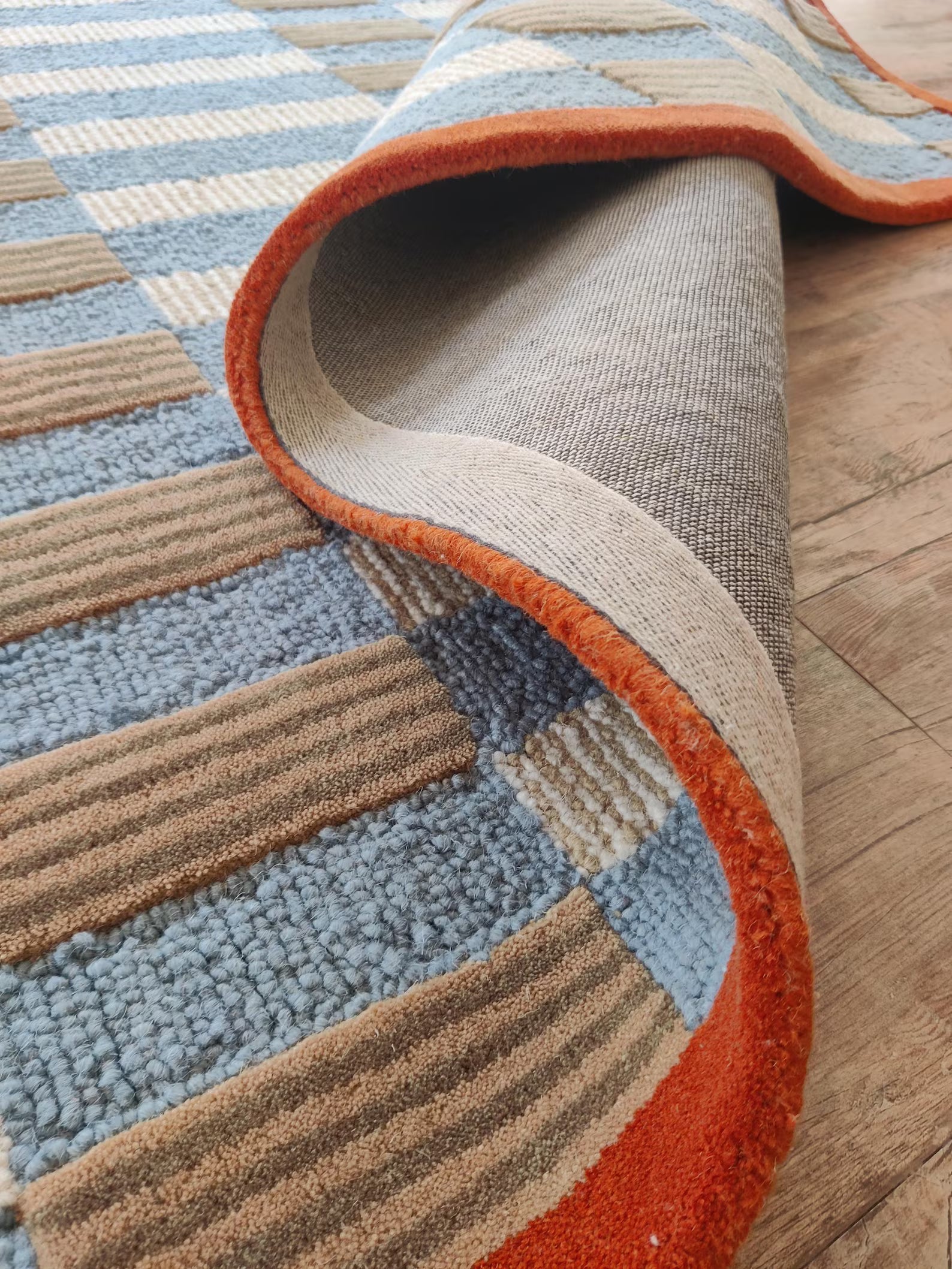 A close-up of a striped area rug with a folded corner showing the underside, set on a light wood floor. The rug features alternating stripes of light blue, tan, and cream, finished with a vibrant orange border.