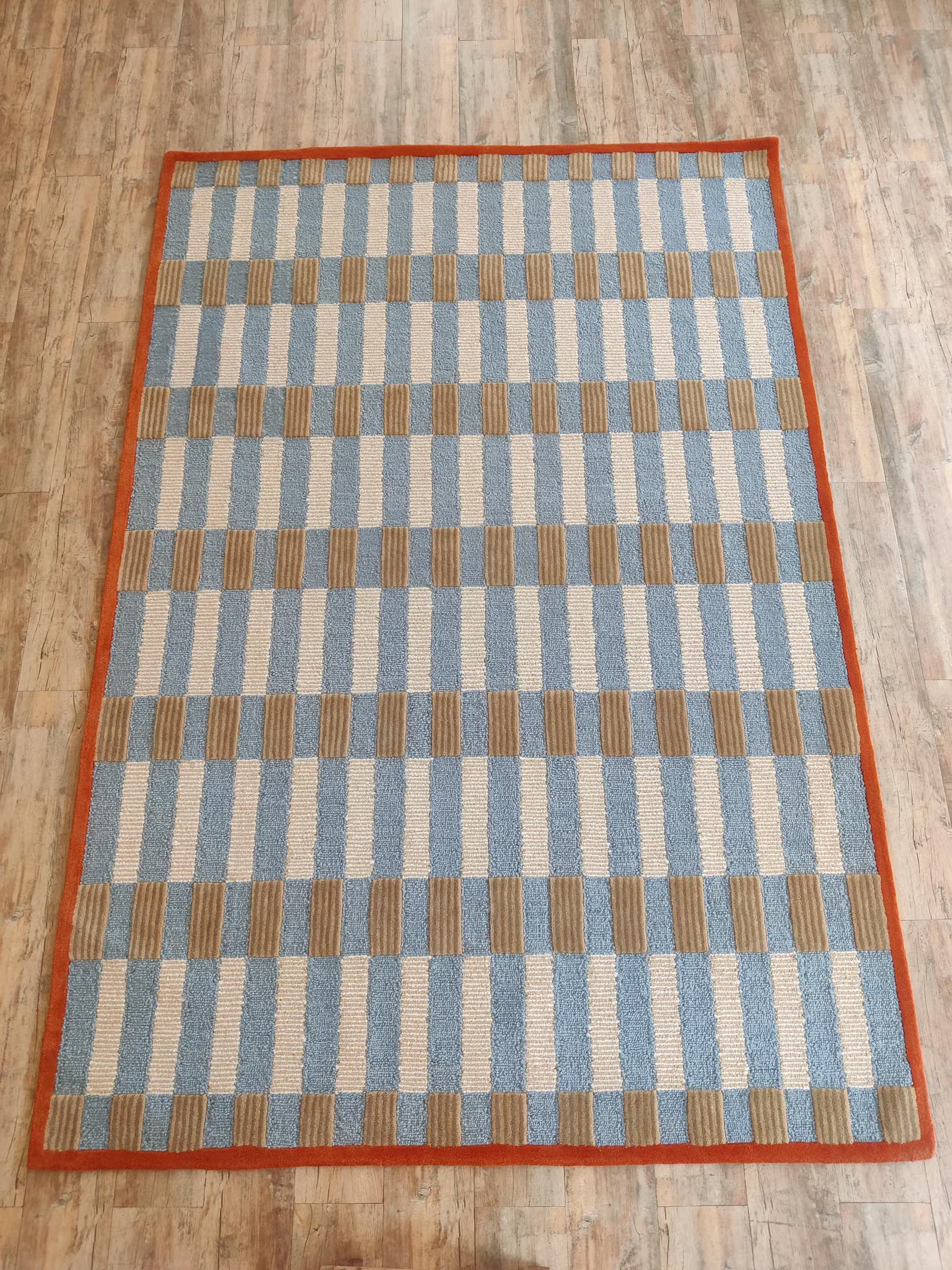 A rectangular area rug with a geometric pattern of light blue, beige, and brown stripes, finished with a solid orange border, shown on a light wood floor.
