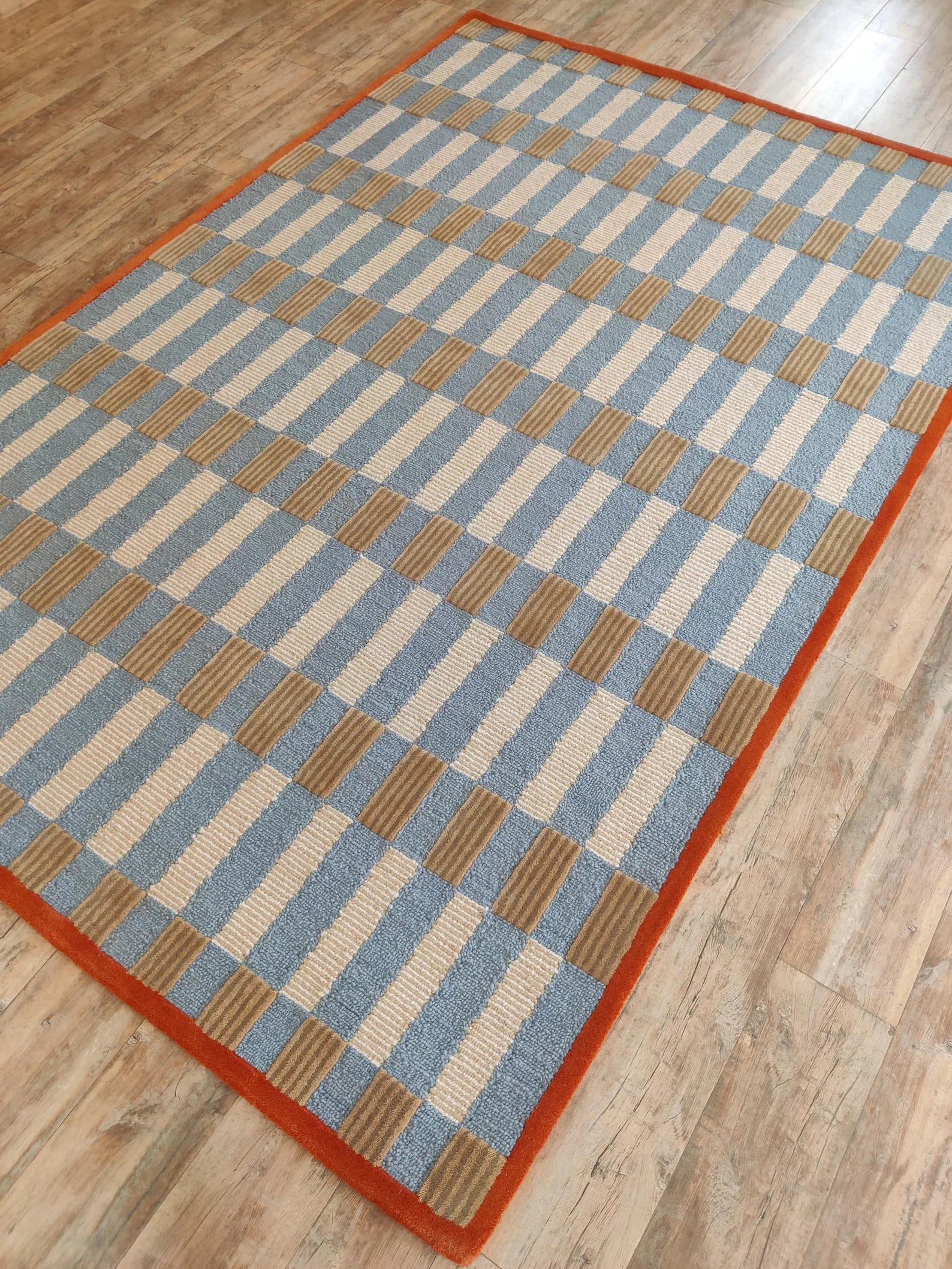 A rectangular area rug with a blue, beige, and brown checkered pattern and a solid orange border, shown on a light wood floor.