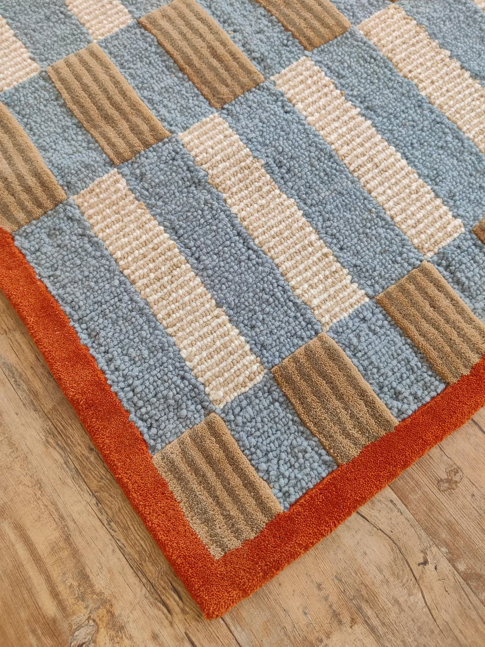 A close-up of a textured area rug with a geometric patchwork design in light blue, beige, and brown, finished with a burnt orange border, shown on a light wood floor.