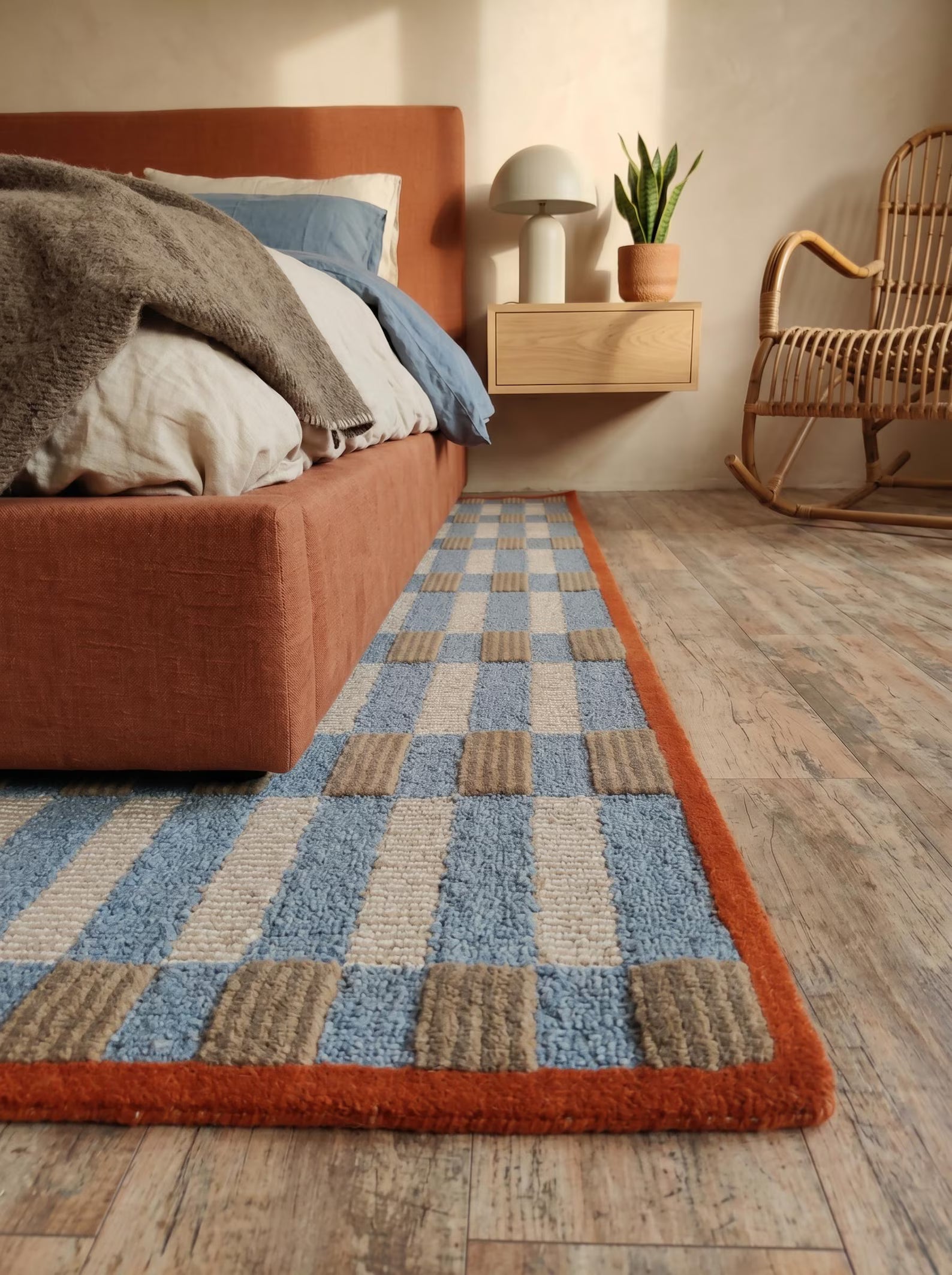 A rectangular area rug with a blue, beige, and brown checkered pattern and a rust-orange border, styled in a cozy bedroom setting next to a bed and a rocking chair.