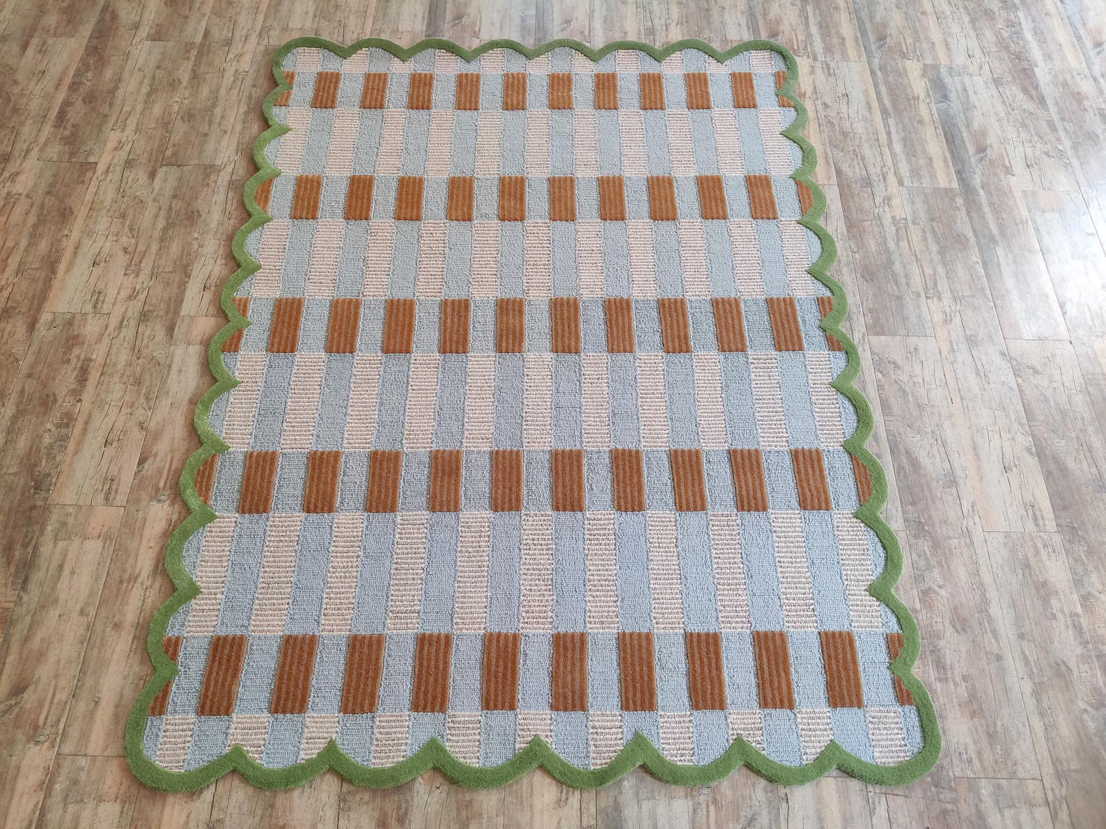 A rectangular woven area rug with a scalloped green border, featuring a checkered pattern of light blue, natural beige, and brown stripes, displayed on a light wood floor.