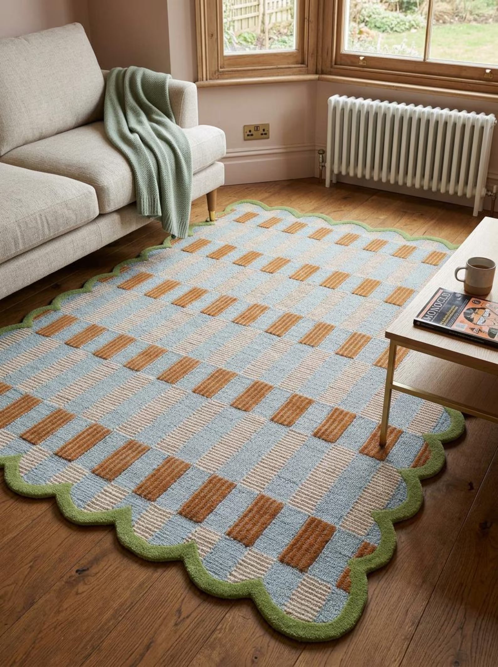 A large rectangular area rug with a scalloped green border, featuring a retro checkerboard pattern in light blue, beige, and brown, styled in a cozy living room on a wooden floor.
