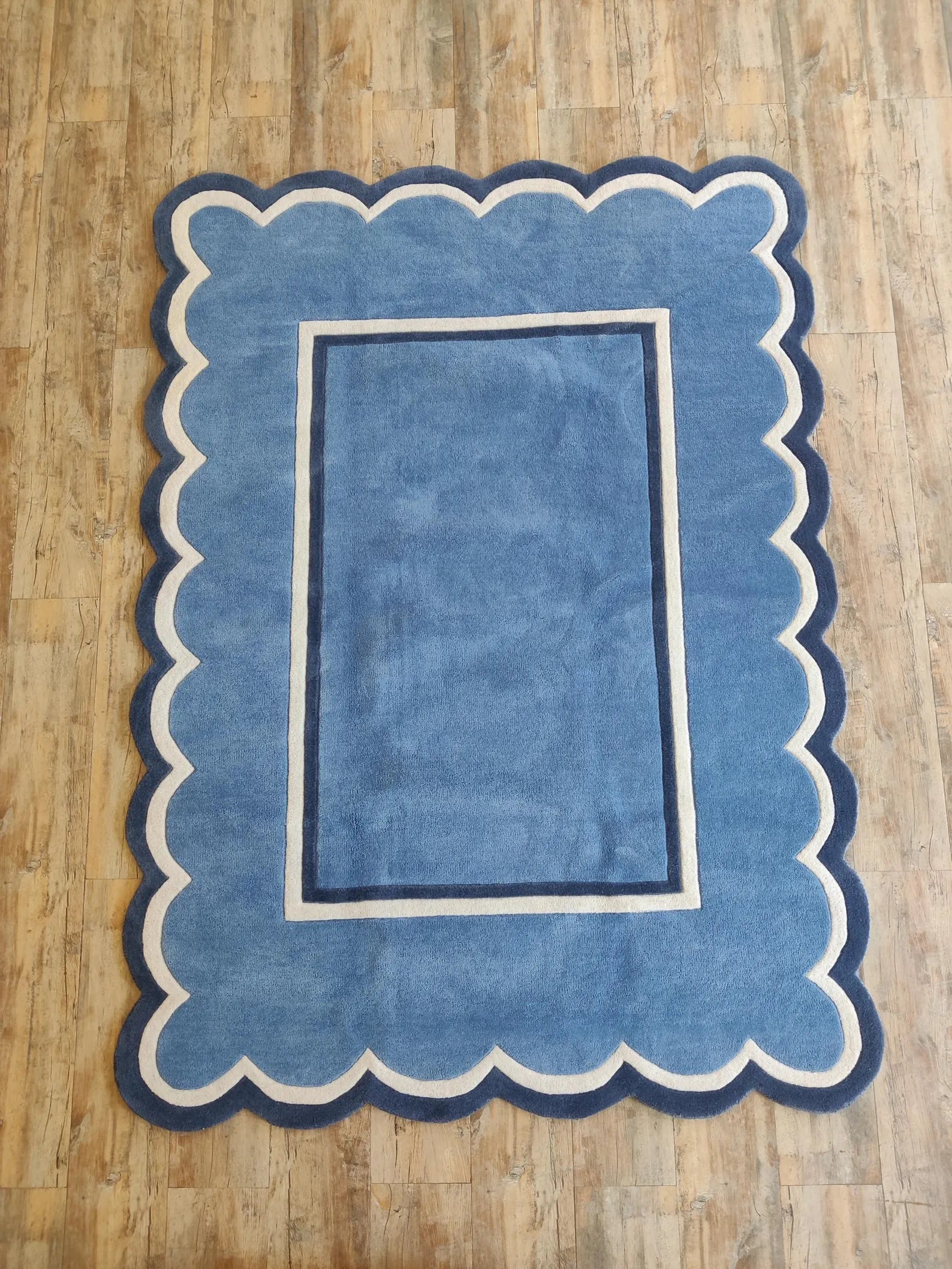 A rectangular blue rug with a scalloped border and a double-line inner border, laid flat on a light brown wooden floor.