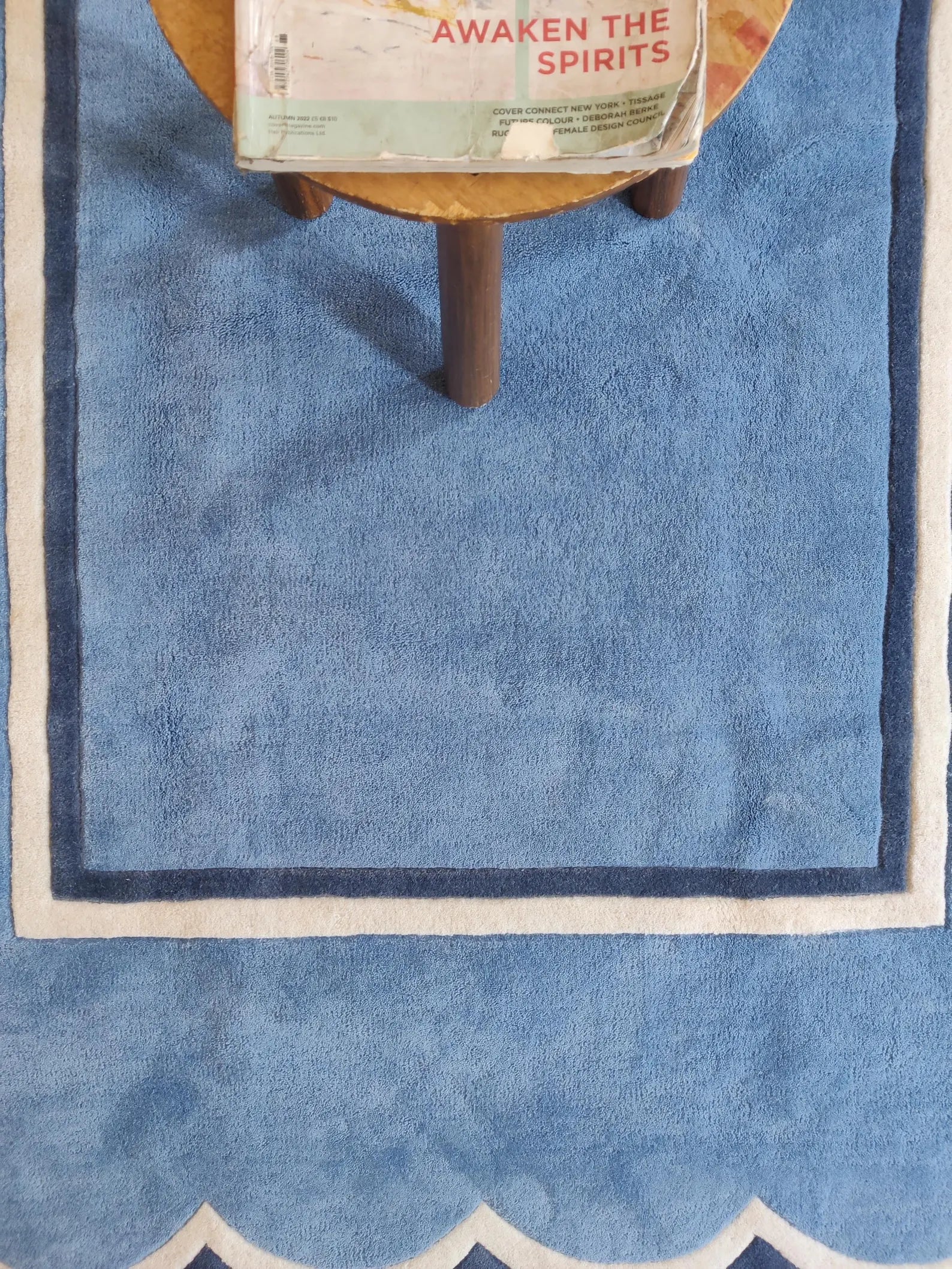 A blue rectangular rug with a white and dark blue border and scalloped edge, shown with a small wooden stool and a book on top.