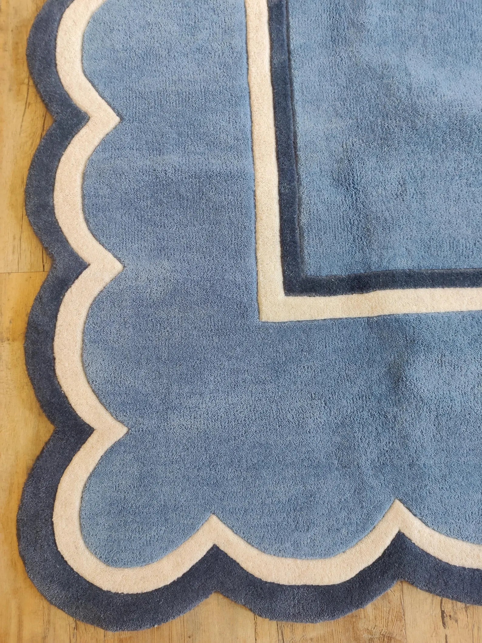 A close-up of a blue rug with a scalloped edge and cream-colored borders, laid on a light wood floor.