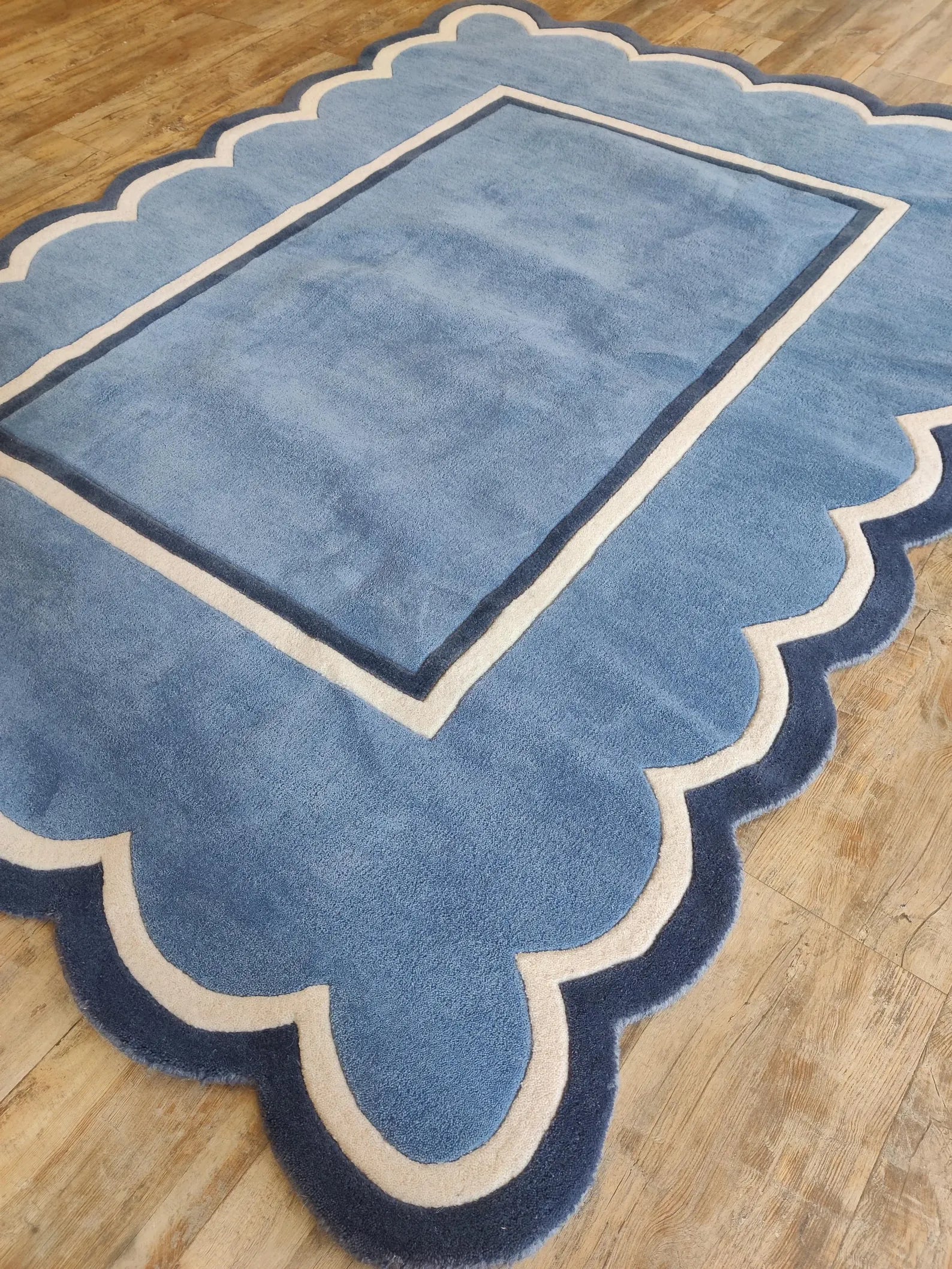 A blue area rug with a scalloped border and a central rectangular panel, shown on a light wood floor.