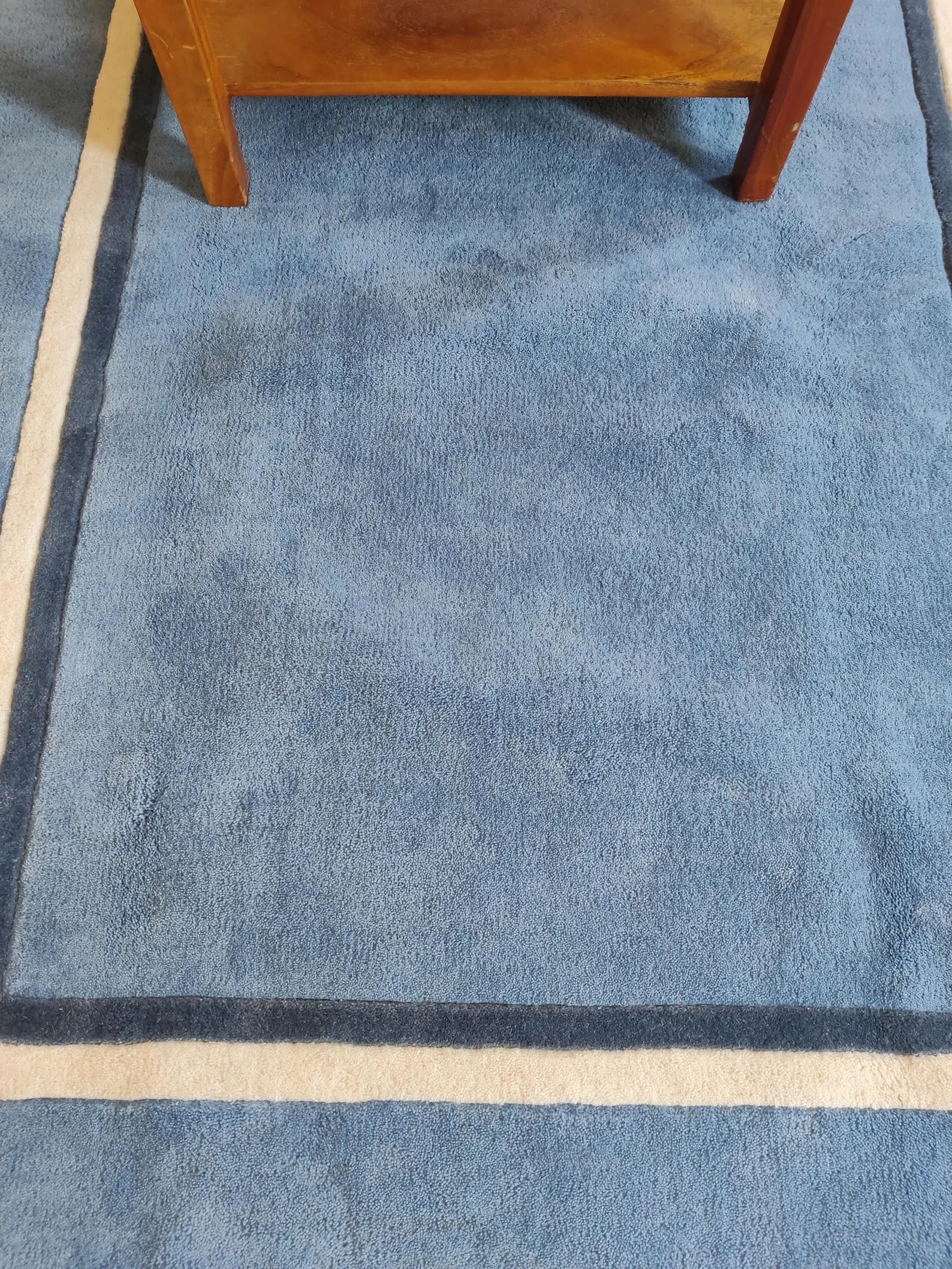 A close-up of a blue textured rug with dark navy and cream borders, with the lower legs of a wooden chair visible at the top edge.