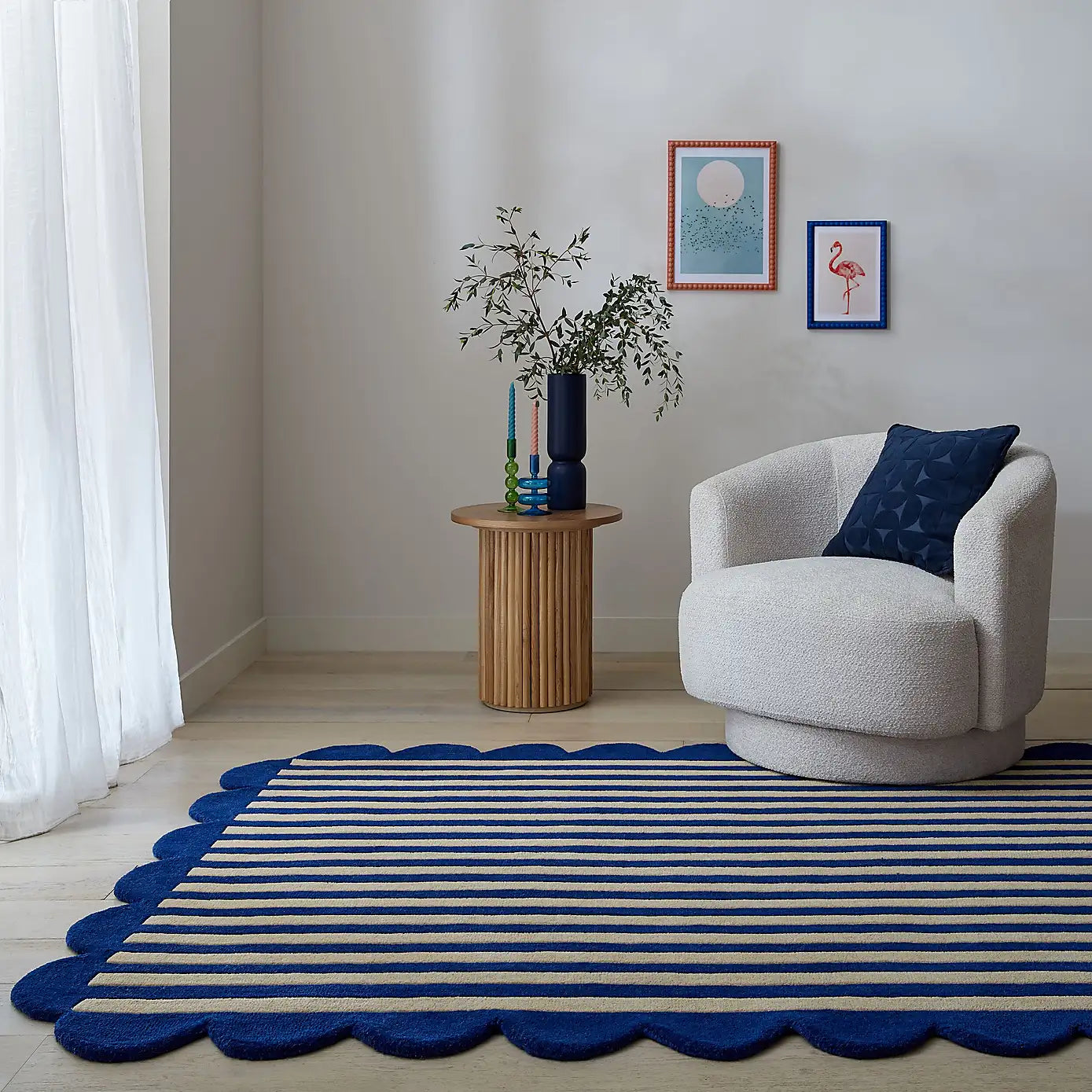A styled interior scene showing a blue and beige striped rug with a scalloped edge in a modern living room corner. A cream upholstered armchair with a dark blue pillow sits beside a small round wooden side table holding a dark blue vase with greenery.