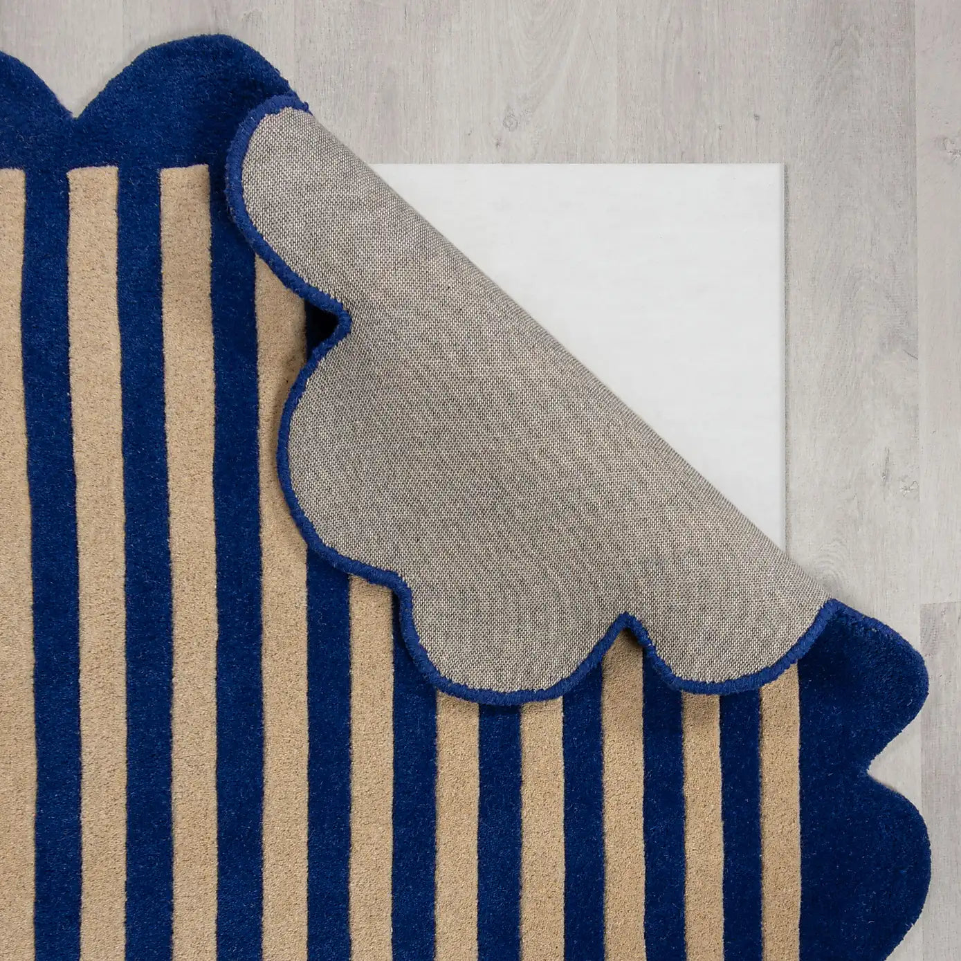A close-up of a striped rug with navy blue and beige vertical stripes, shown with one scalloped corner folded over to reveal the textured beige underside, placed on a light gray wood floor.